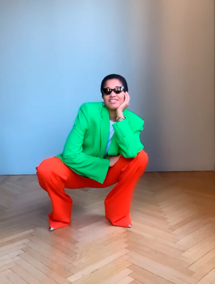 A girl crouched wearing a color mixes: red pants, green blazer and sunglasses