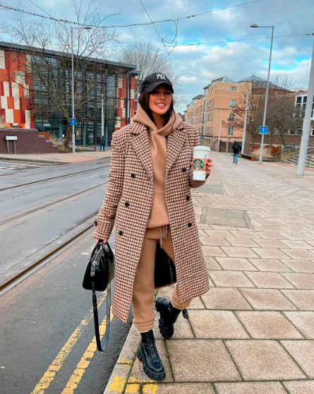 A girl in the street with a cofee wearing a tracksuit with a long coat, a black hand bag and a hat